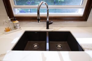 Close-up of double deep kitchen sinks with marble counters, stainless steel faucet, and subway tile backsplash.